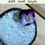 Homemade viola bath salt in a small bowl with a fresh viola flower.