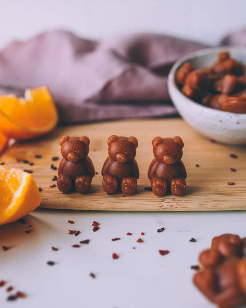 3 vitamin C gummy bears sitting up on a wooden cutting board, with orange slices surrounding.