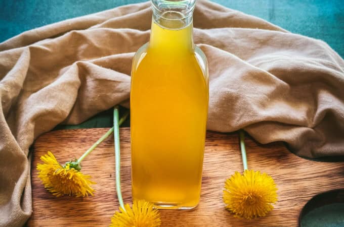A bottle of yellow dandelion syrup on a wood cutting board surrounded by fresh dandelion flowers, natural fabric, and a teal background.