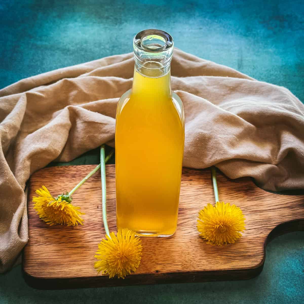 A bottle of yellow dandelion syrup on a wood cutting board surrounded by fresh dandelion flowers, natural fabric, and a teal background. 