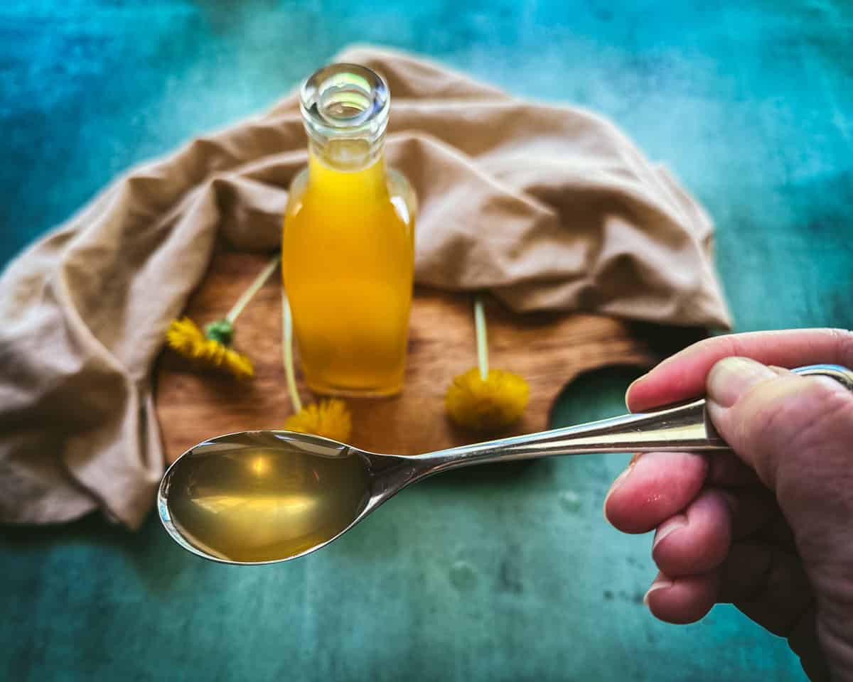 Dandelion syrup on a spoon, lifted from a bottle of dandelion syrup on a natural cloth on a teal background. 