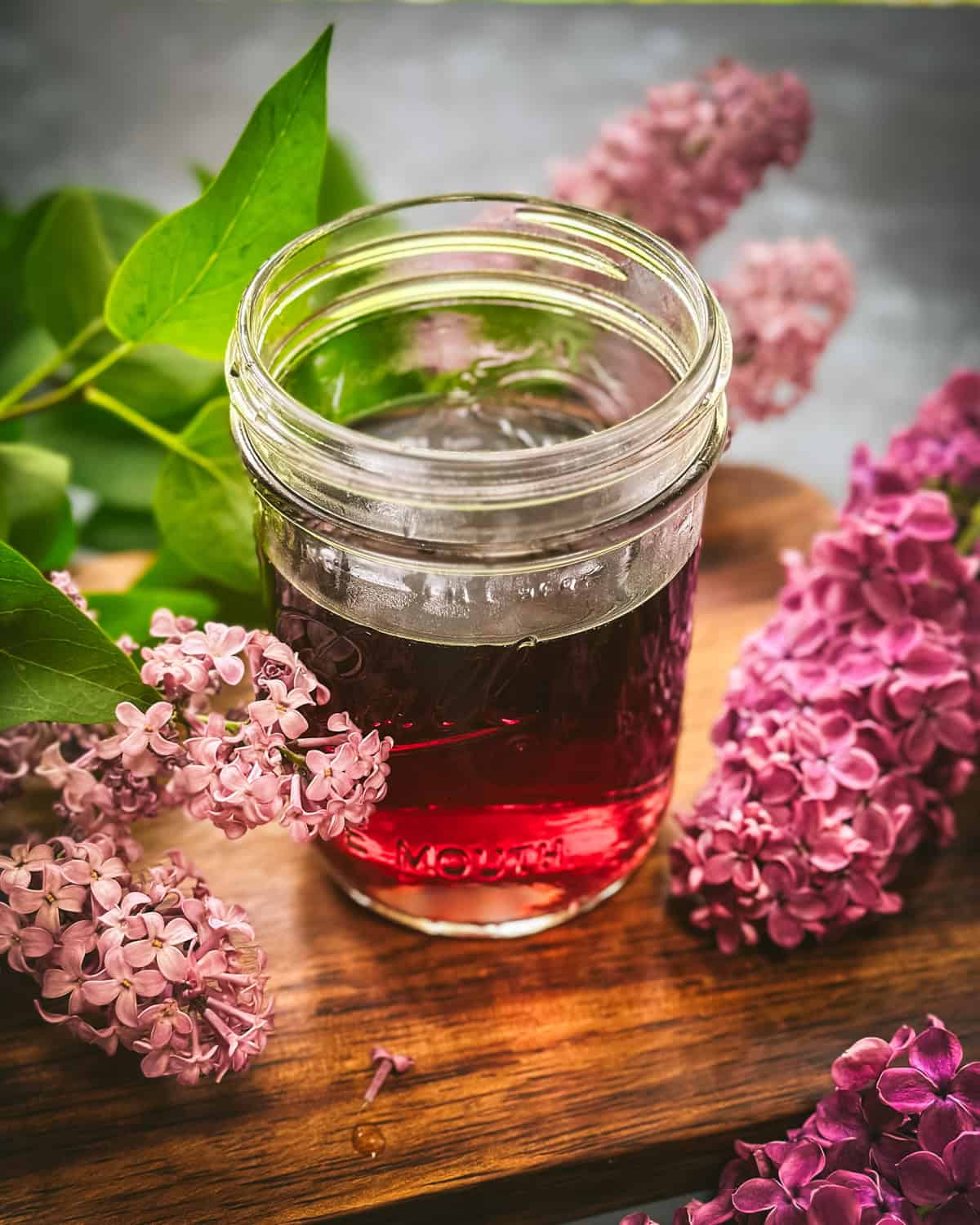 A jar of lilac tea on a wood cutting board surrounded by fresh lilacs. 