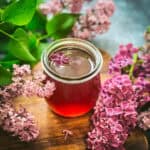 A jar of lilac syrup on a wood cutting board surrounded by fresh lilac flowers and leaves.