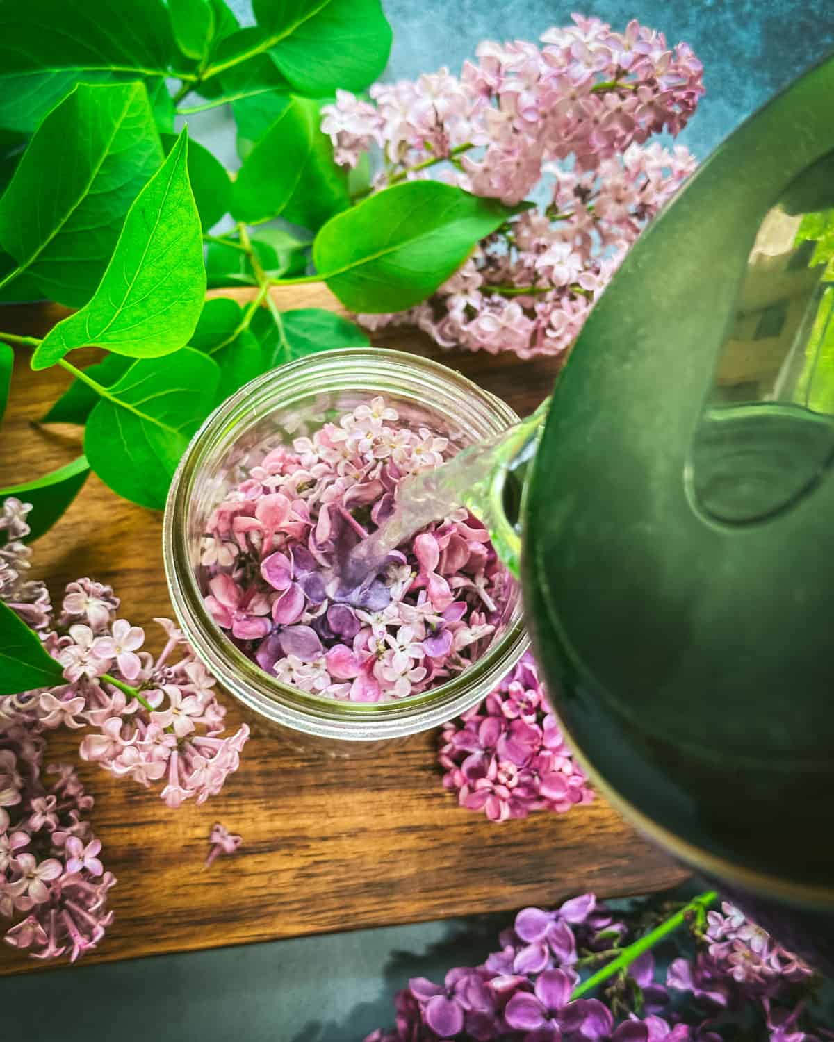 Boiling water pouring from a green kettle into a jar of lilac petals, surrounded by fresh lilacs. 