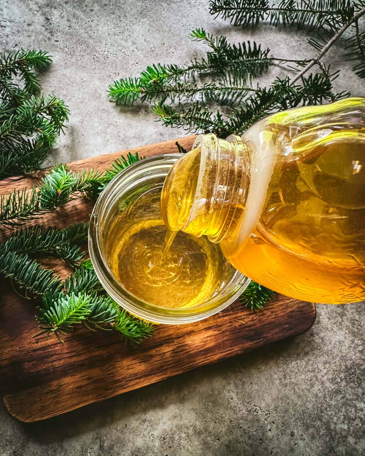 Honey pouring into the fir tip tea, on a wood cutting board surrounded by fresh fir tips. 