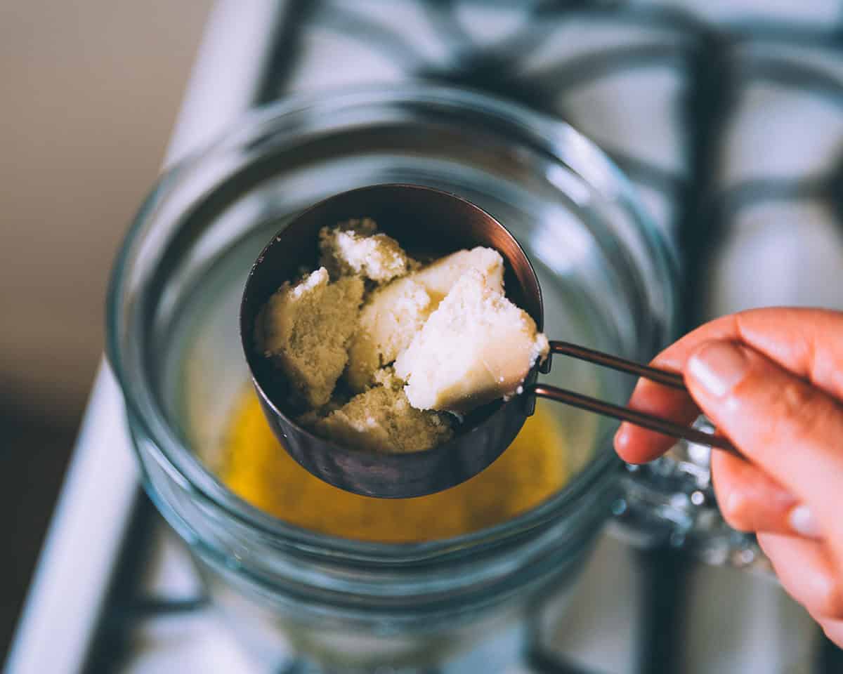 A measuring cup with shea butter pouring into the top of a double boiler on a gas stove. 