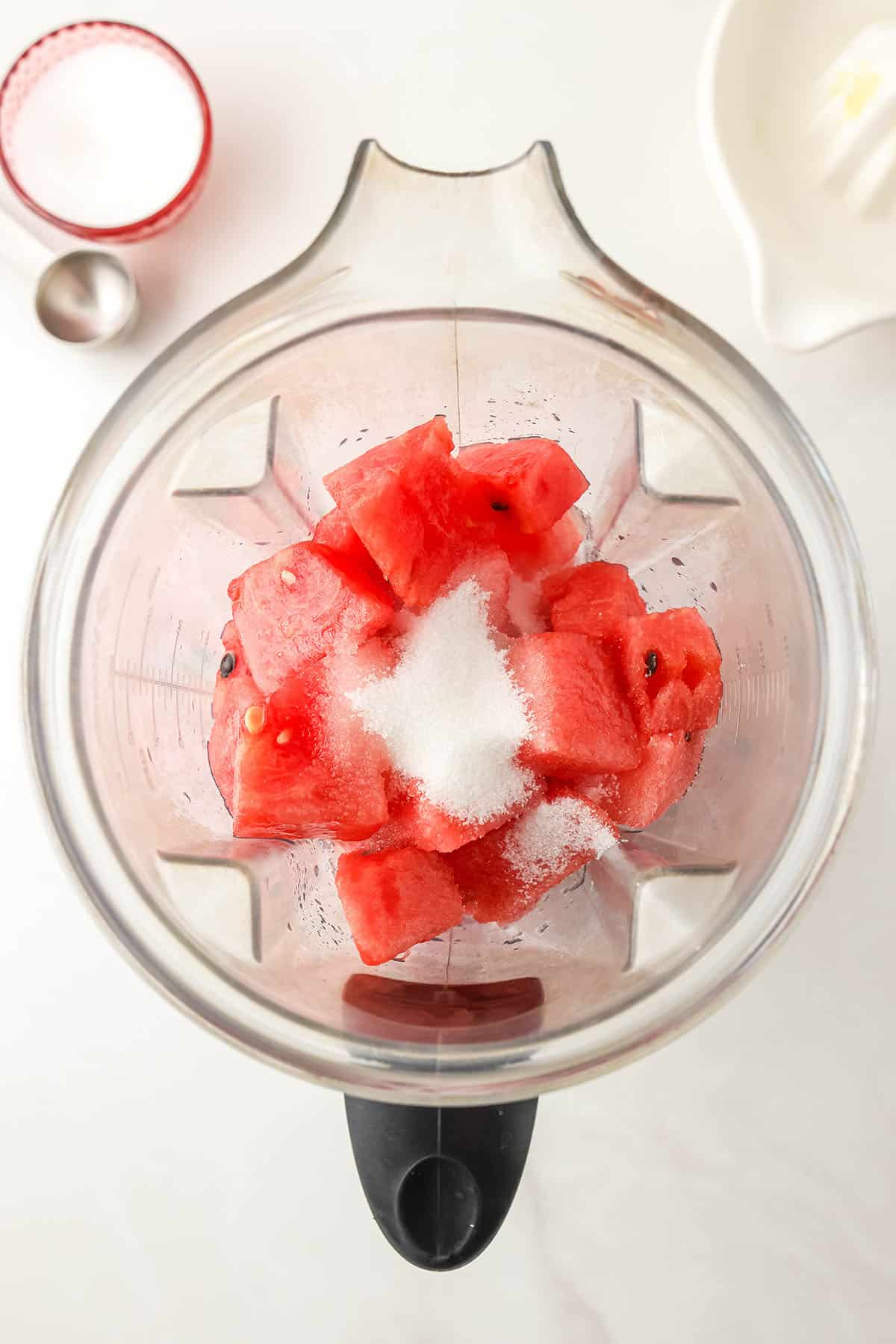 Cubed watermelon and sugar in a blender on a white countertop, top view. 