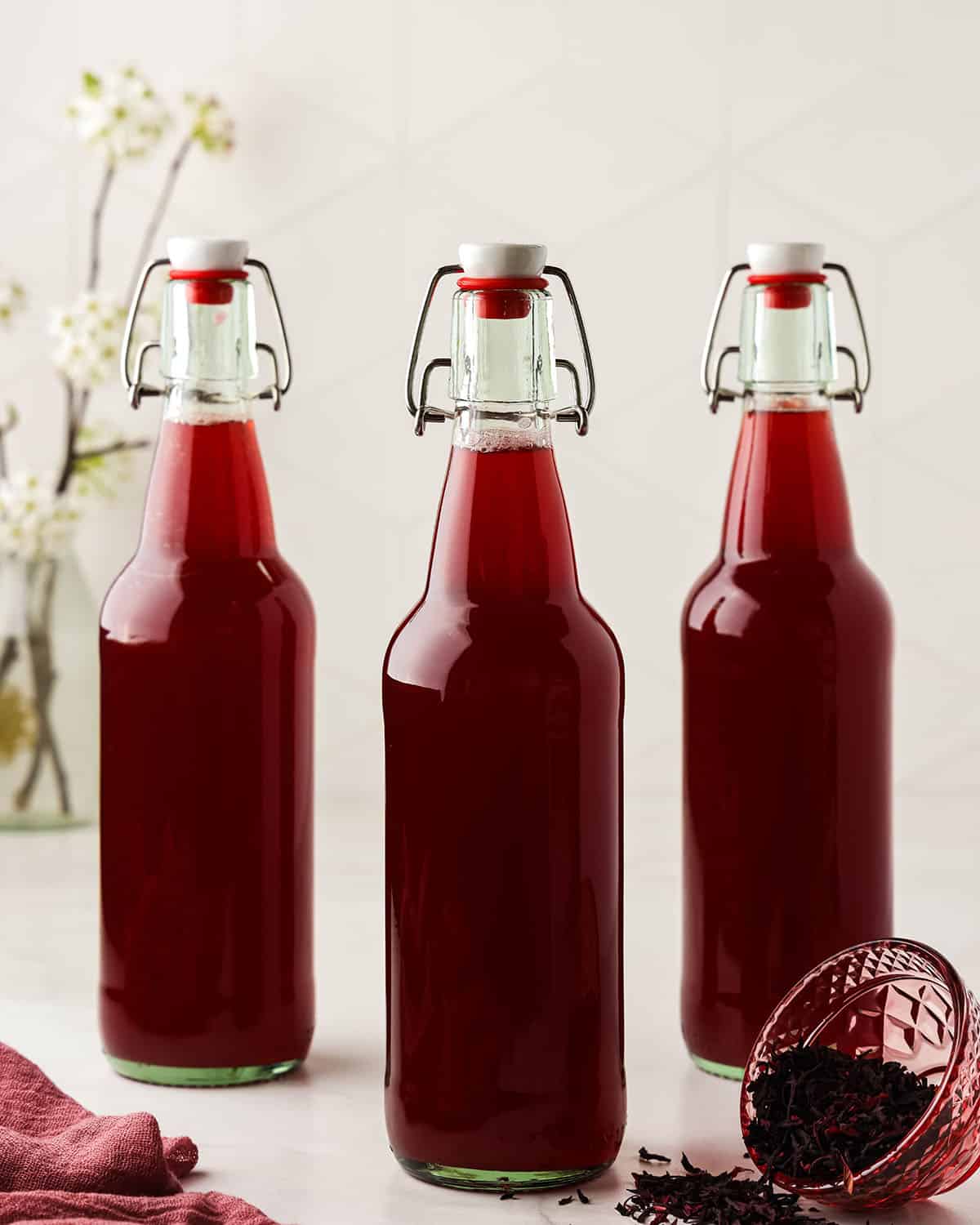 3 bottles filled with hibiscus tea and kombucha, surrounded by loose hibiscus tea. 
