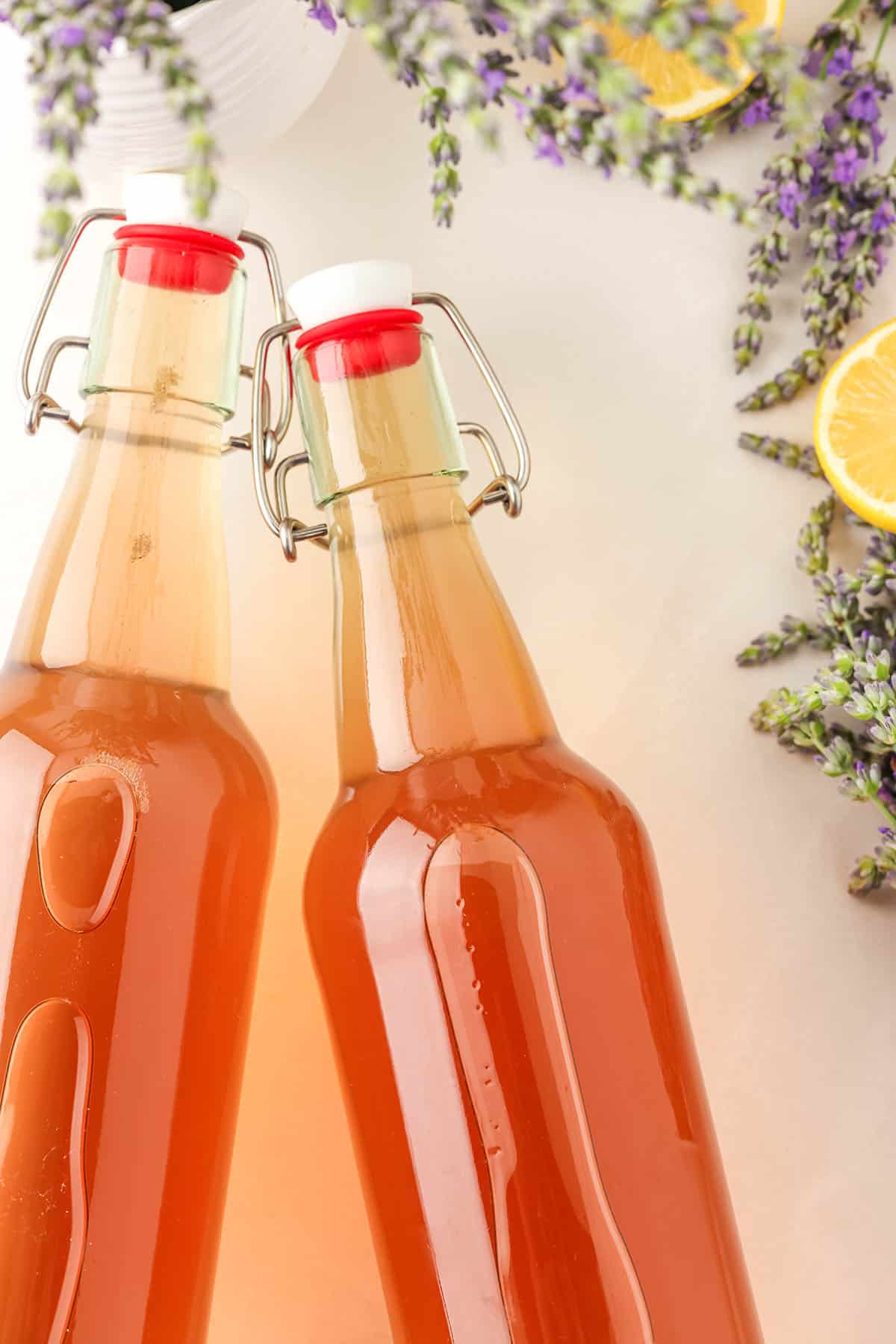 2 sealed bottles of lavender kombucha on their sides on a white countertop surrounded by fresh lemons and lavender. 