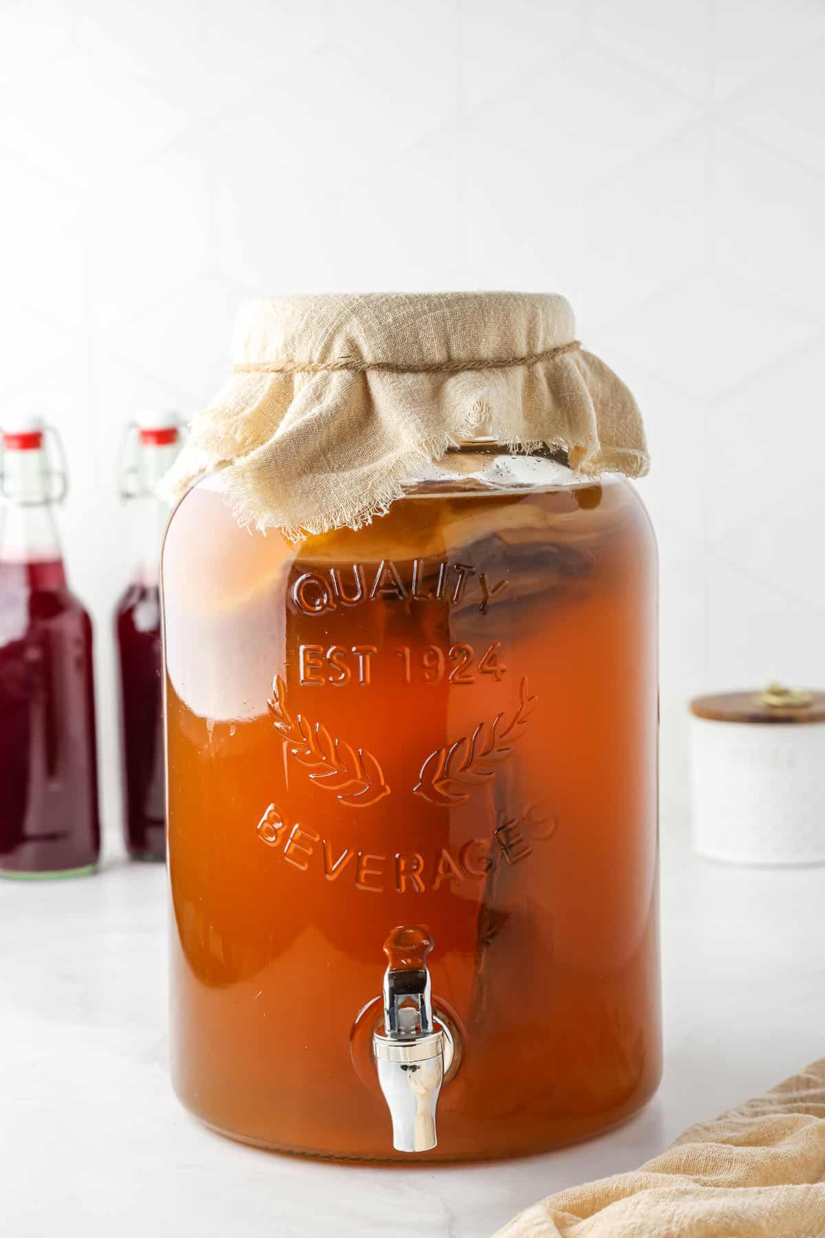 A wide mouth gallon jar with green tea and a SCOBY floating on top, covered with cheesecloth and secured with a rubber band. 