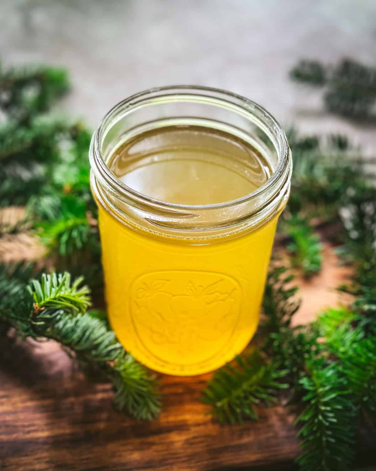 A jar of golden colored fir tip syrup, on a wood cutting board surrounded by fresh fir tips. 