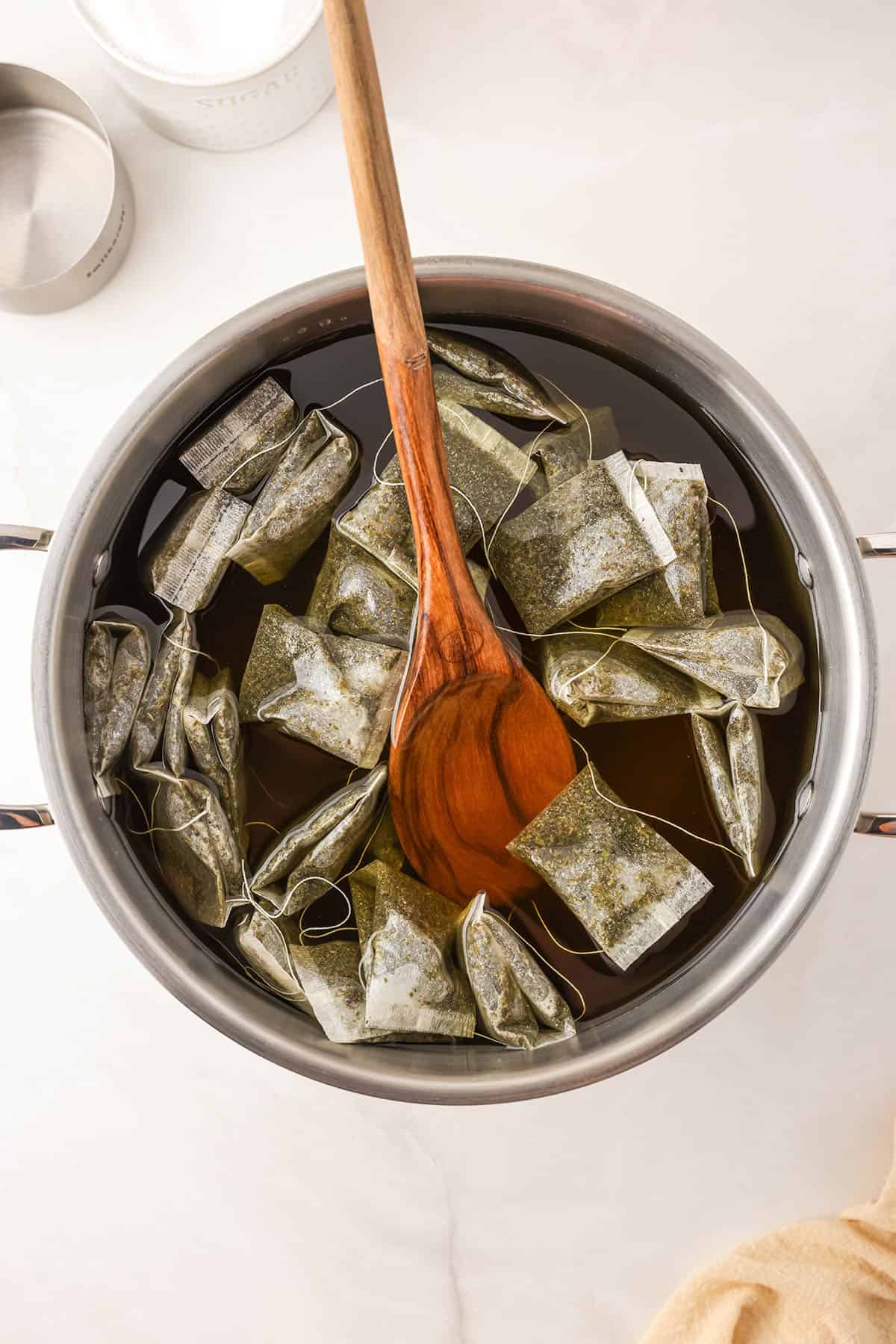 A large pot with water and several tea bags brewing, with a wooden spoon rested into the water and stirring. 