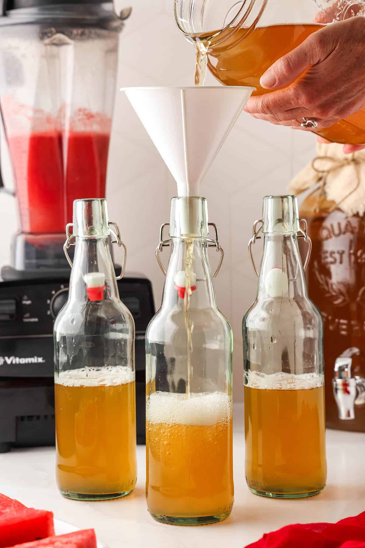 Swing top bottles with a funnel on one that has kombucha pouring into it, the bottles halfway full. A blender with watermelon puree is in the background. 