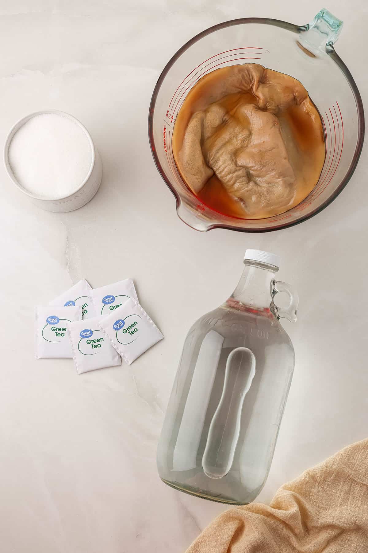 A glass bottle of water on its side, green tea bags, a scoby in a glass pyrex, and sugar in a dish, top view. 