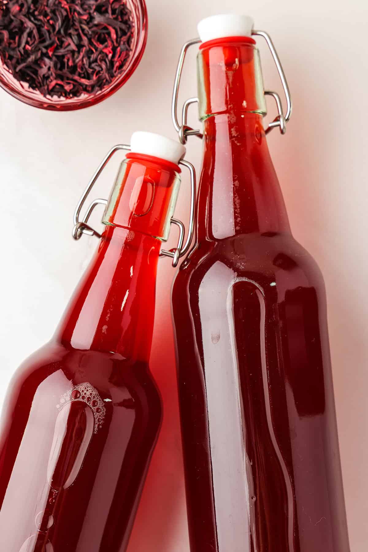 2 bottles filled with hibiscus kombucha, with a small pink bowl filled with loose hibiscus tea to the upper left. 