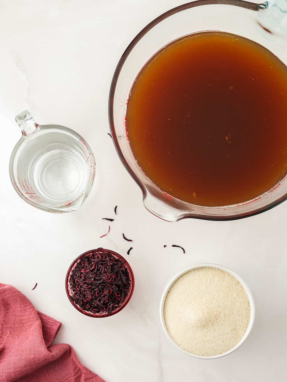 Ingredients to make hibiscus kombucha in bowls, top view. On a white countertop with dried tea and a dark pink cloth napkin surrounding. 