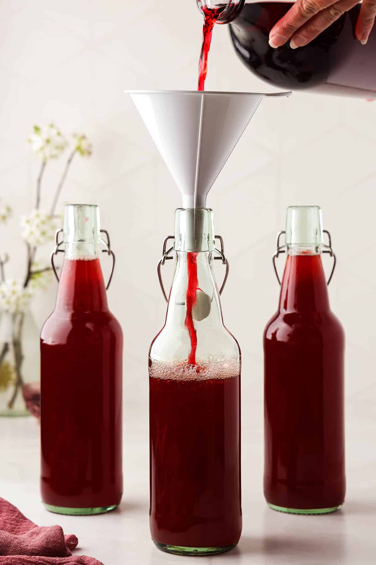 3 flip top bottles with kombucha topped off with hibiscus tea, the front bottle still being filled with a funnel on top. 