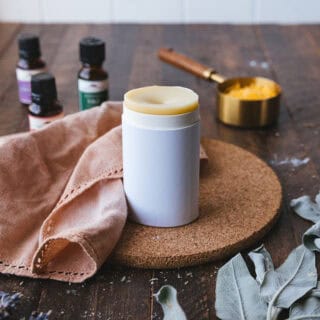 A homemade deodorant stick in a cardboard container, on a cork circular board. Surrounded by a natural cloth napkin, sage leaves, and essential oils on a dark wood surface.