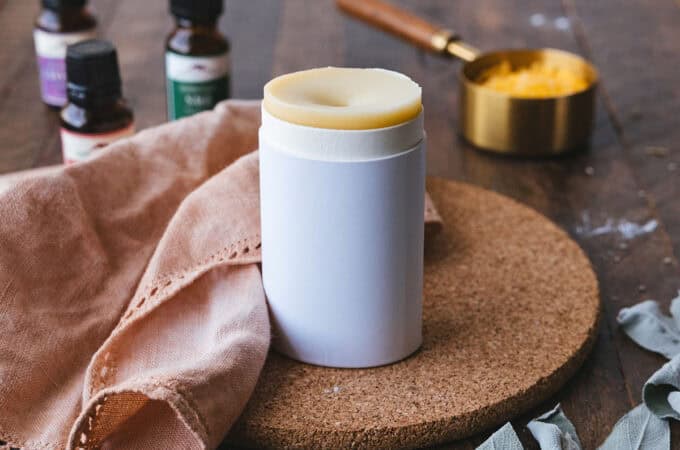 A homemade deodorant stick in a cardboard container, on a cork circular board. Surrounded by a natural cloth napkin, sage leaves, and essential oils on a dark wood surface.