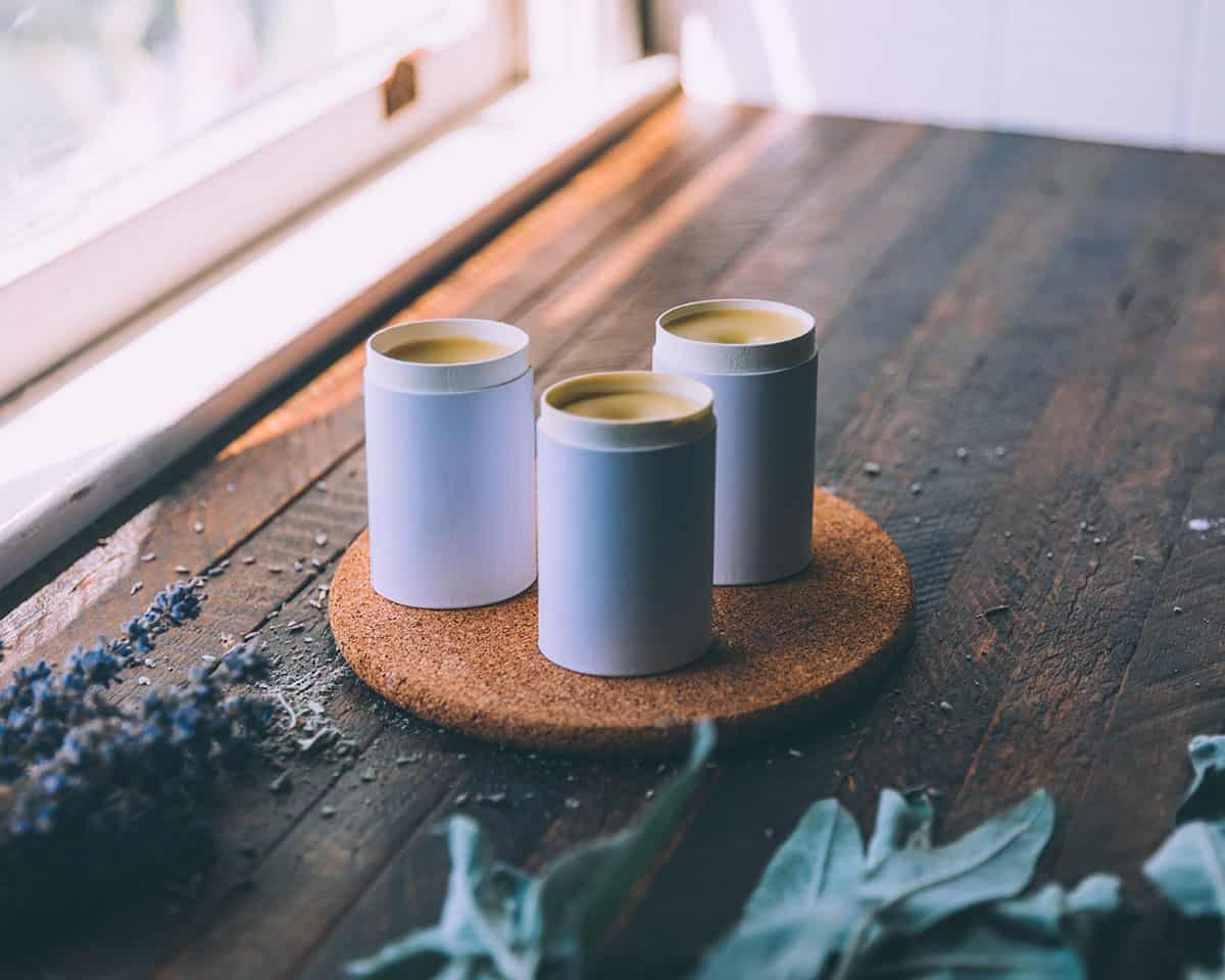 3 cardboard tubes of deodorant setting up on a circle cork board, on a dark wood surface with herbs, in natural light. 