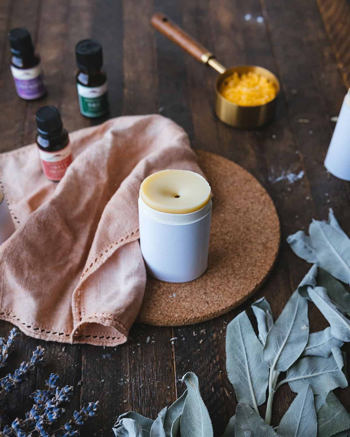 Homemade deodorant in a cardboard tube surrounded with a natural cloth napkin and essential oils, sage leaves, and other ingredients on a dark wood surface.