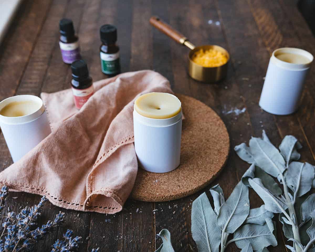 Homemade deodorant in a cardboard tube surrounded with a natural cloth napkin and essential oils, sage leaves, and other ingredients on a dark wood surface.