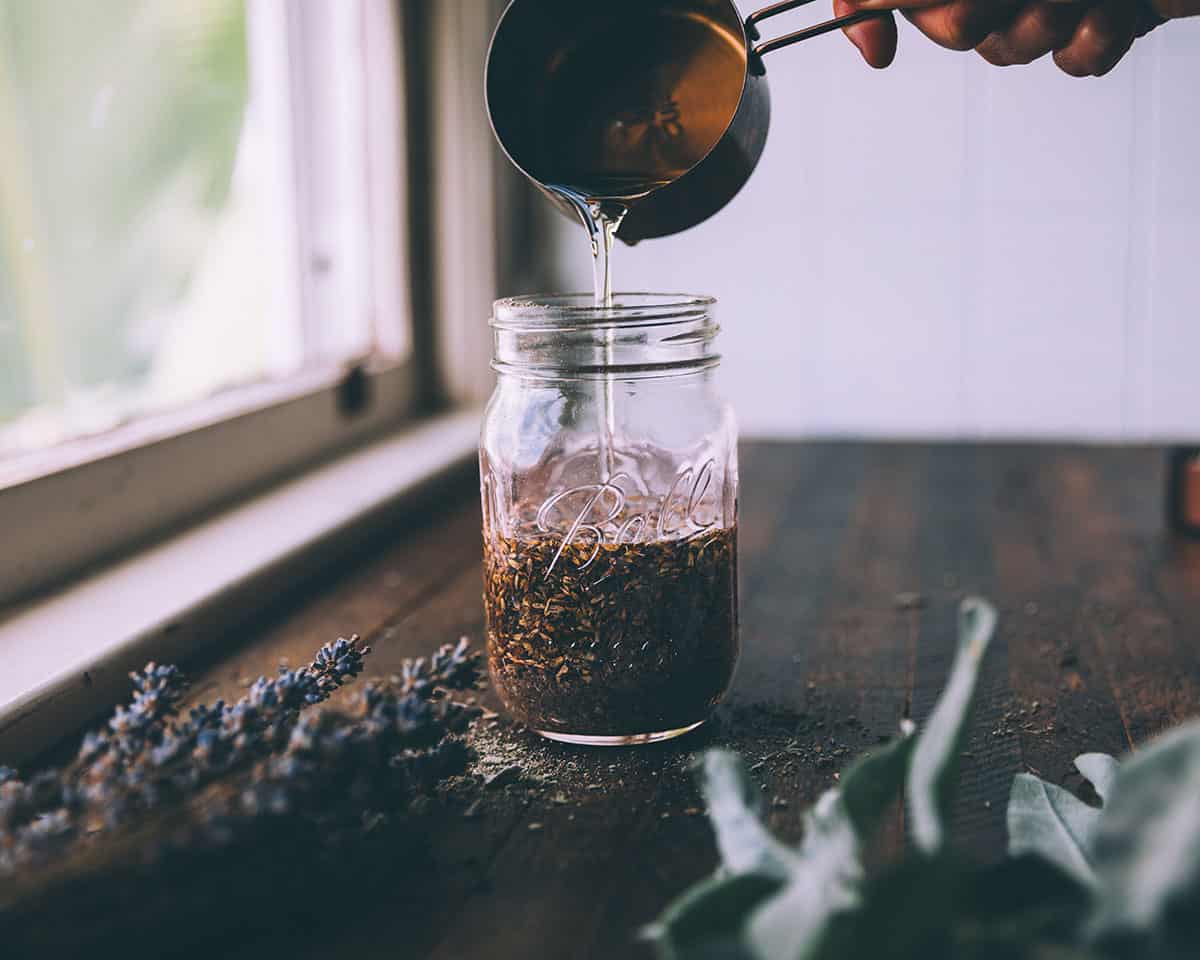 Oil pouring into a jar with dried herbs in it, on a dark wood surface surrounded by dried lavender and sage, with a window in the background. 