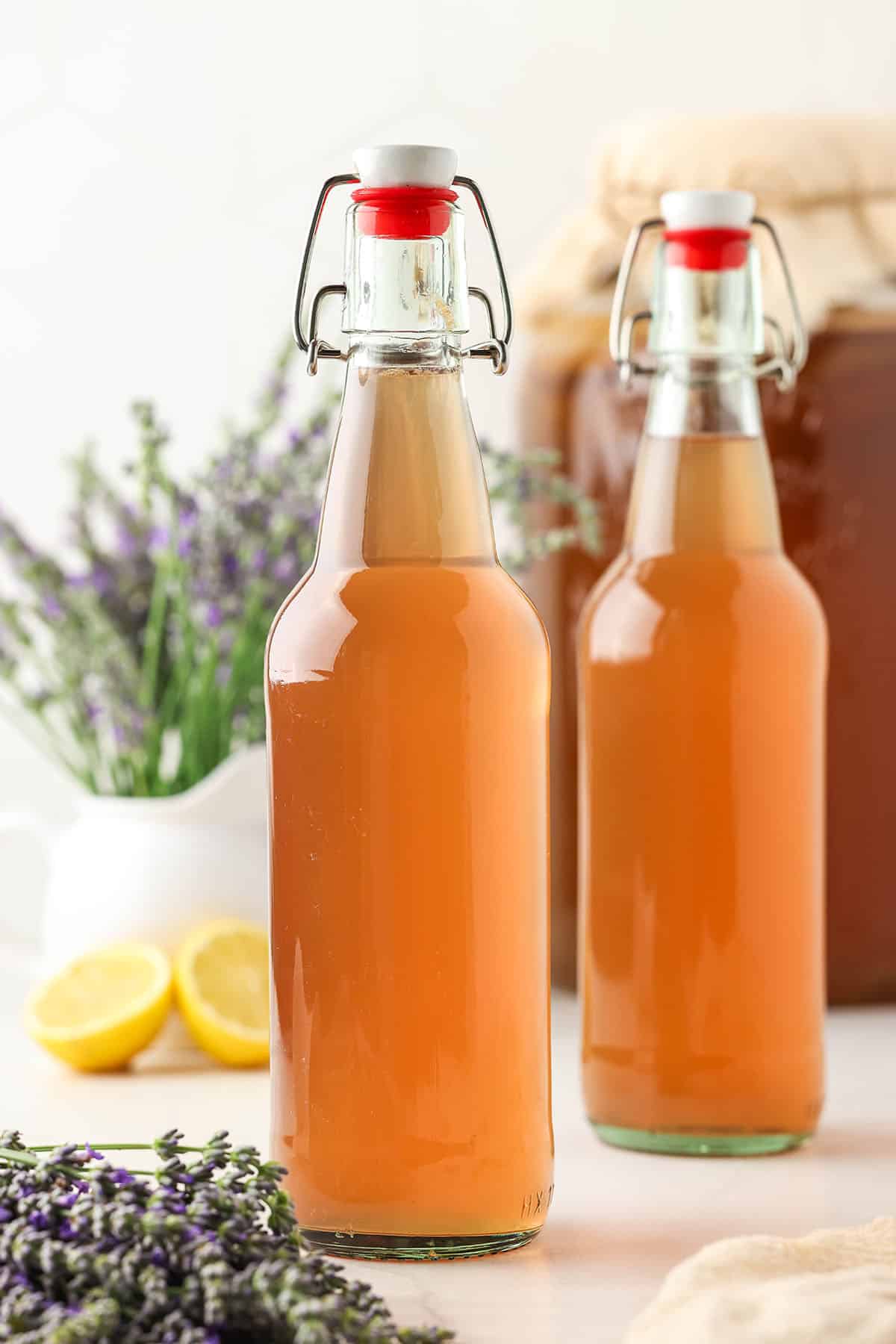 Bottles of lavender kombucha on a white surface surrounded by fresh lavender and lemons.