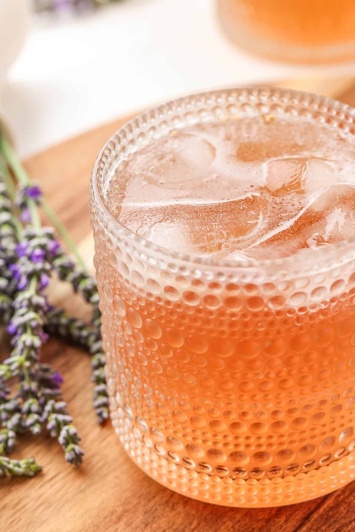 A collins glass of lavender kombucha with ice, on a wood surface with fresh lavender flowers surrounding. 