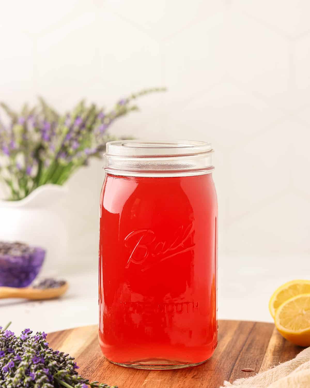 Lavender syrup in a jar, on a wood surface surrounded by fresh lavender flowers and lemons.