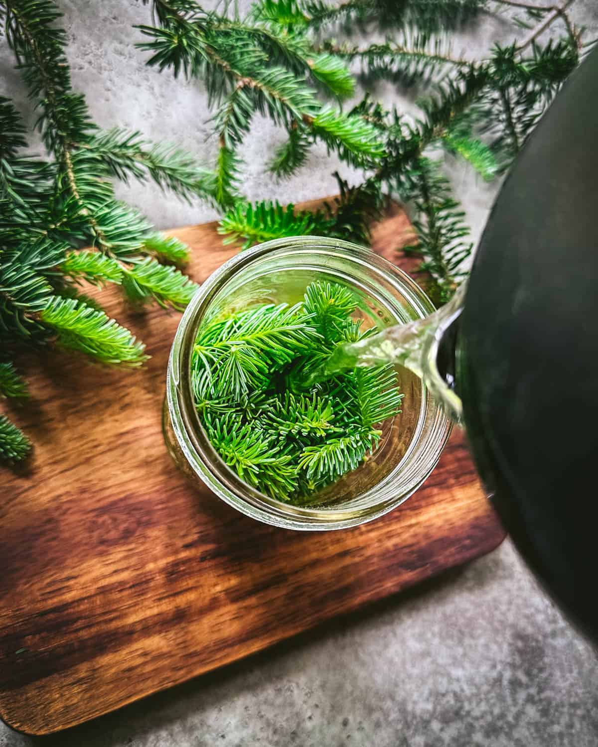 Boiling water pouring into the jar over the fir tips, on a wood cutting board with fir branches surrounding. Top view. 