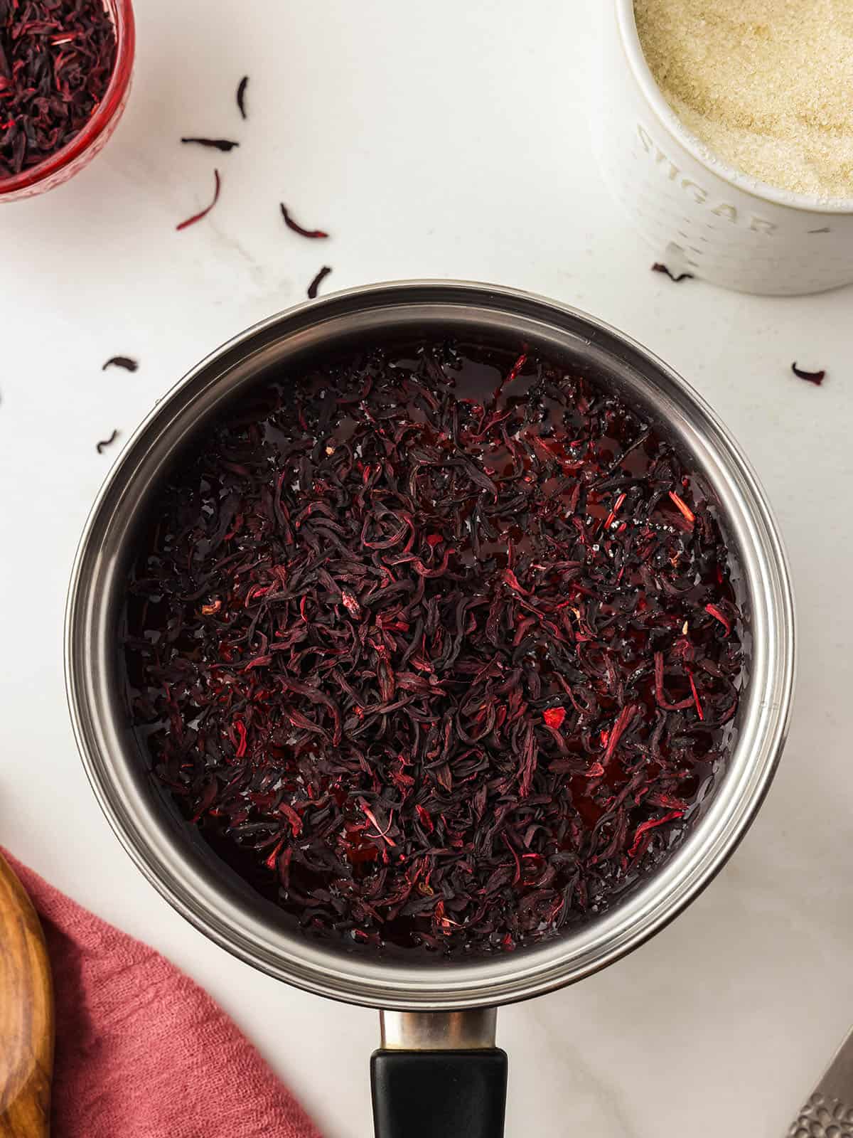 Hibiscus tea in a pot, on a white surface surrounded by loose hibiscus tea. 