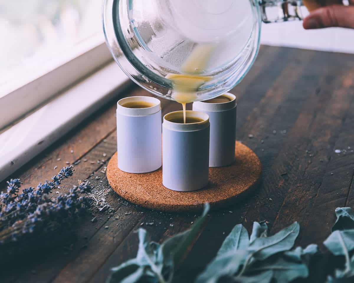 A glass bowl pouring the liquid deodorant into circular cardboard deodorant tubes sitting on a round cork surface, on a dark wood countertop surrounded by herbs. 