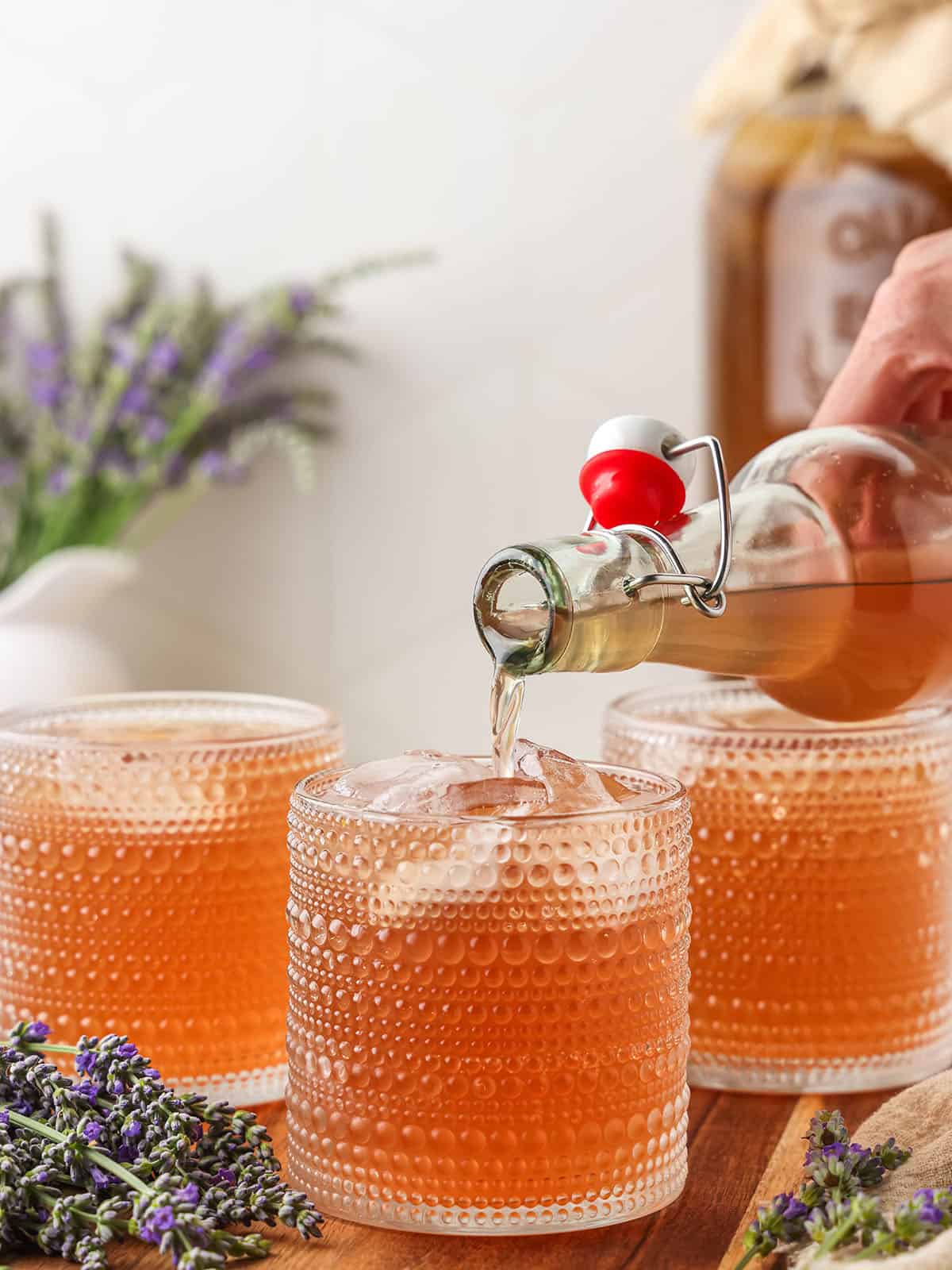 A bottle of lavender kombucha pouring into vintage collins glasses with ice, with fresh lavender flowers in the background. 