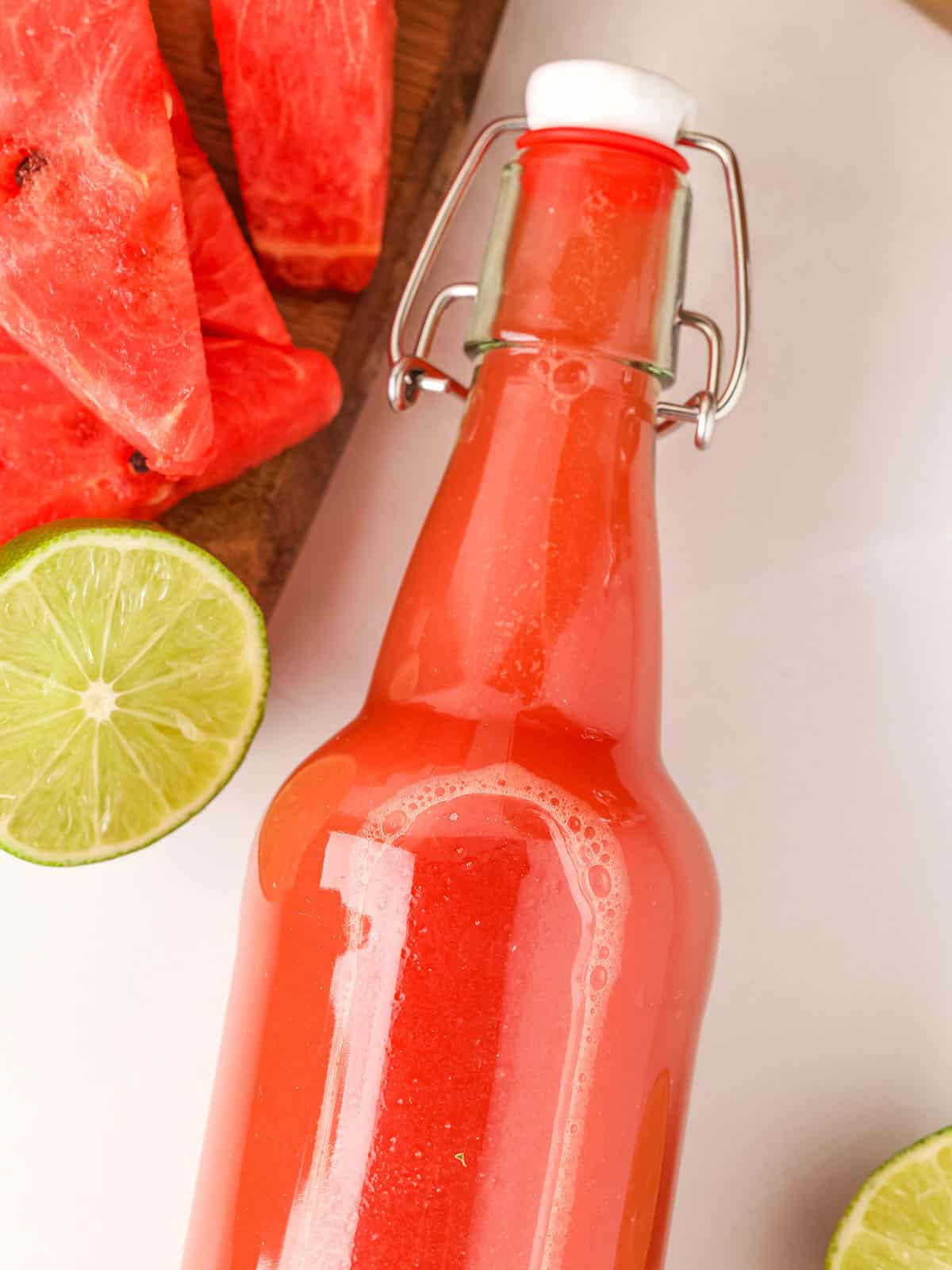 A bottle of watermelon kombucha on its side, with lime halves and sliced watermelon surrounding. 