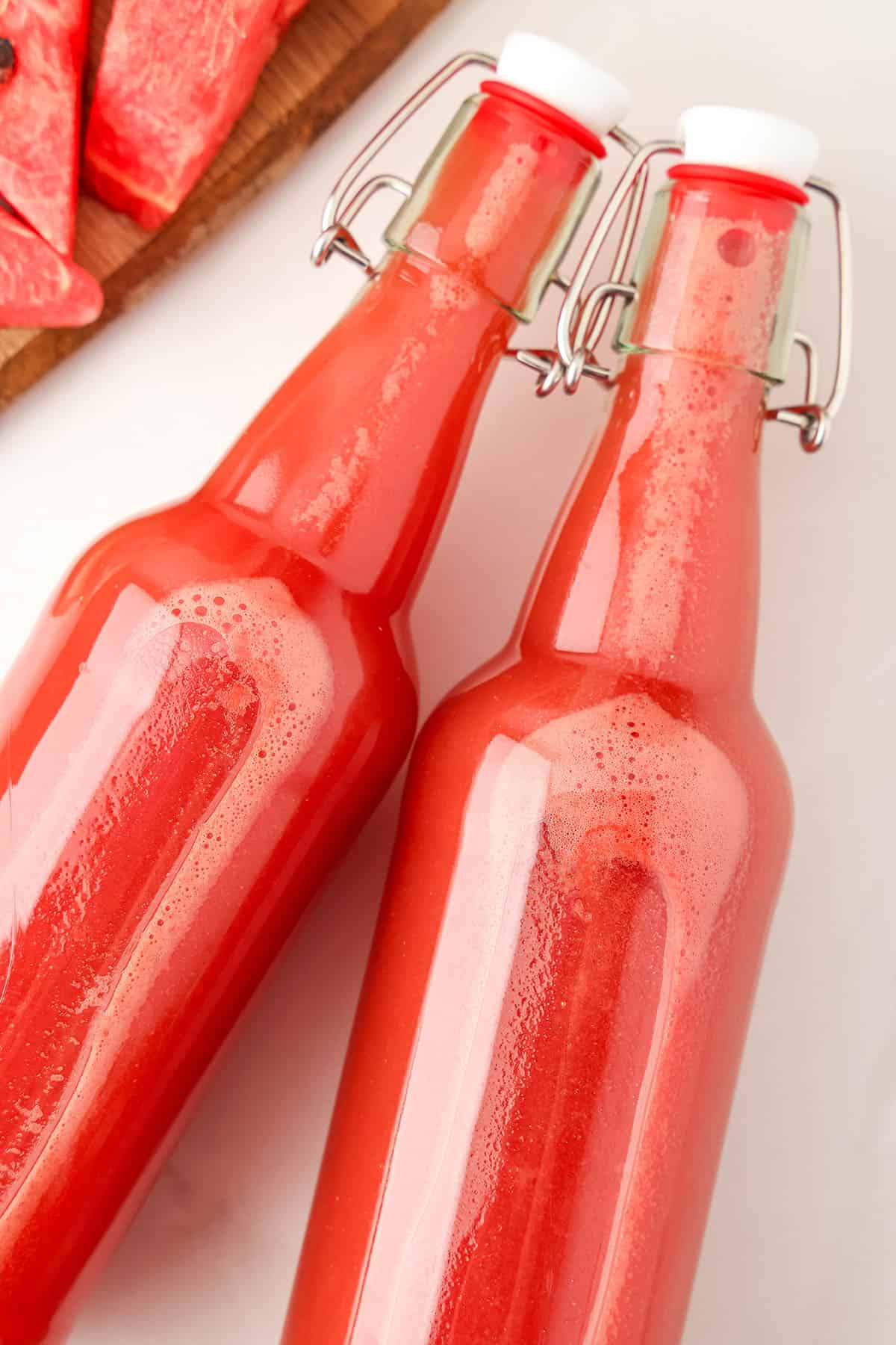 2 bottles of watermelon kombucha on their sides on a white countertop with fresh sliced watermelon to the side. 