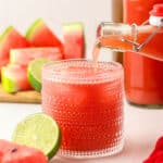 A bottle of homemade watermelon kombucha pouring into a vintage collins glass with ice, surrounded by fresh cut limes and watermelon.