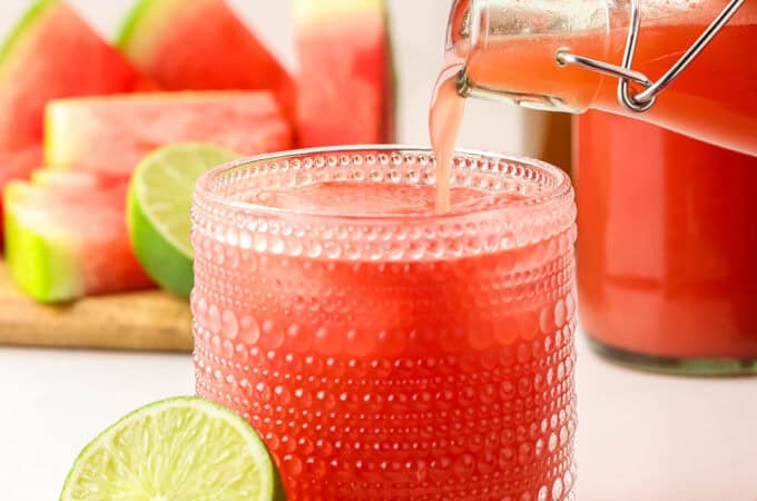 A bottle of homemade watermelon kombucha pouring into a vintage collins glass with ice, surrounded by fresh cut limes and watermelon.
