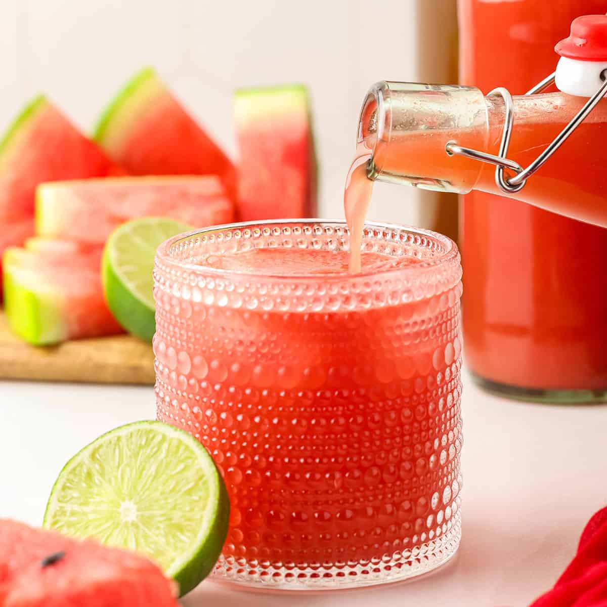 A bottle of homemade watermelon kombucha pouring into a vintage collins glass with ice, surrounded by fresh cut limes and watermelon. 