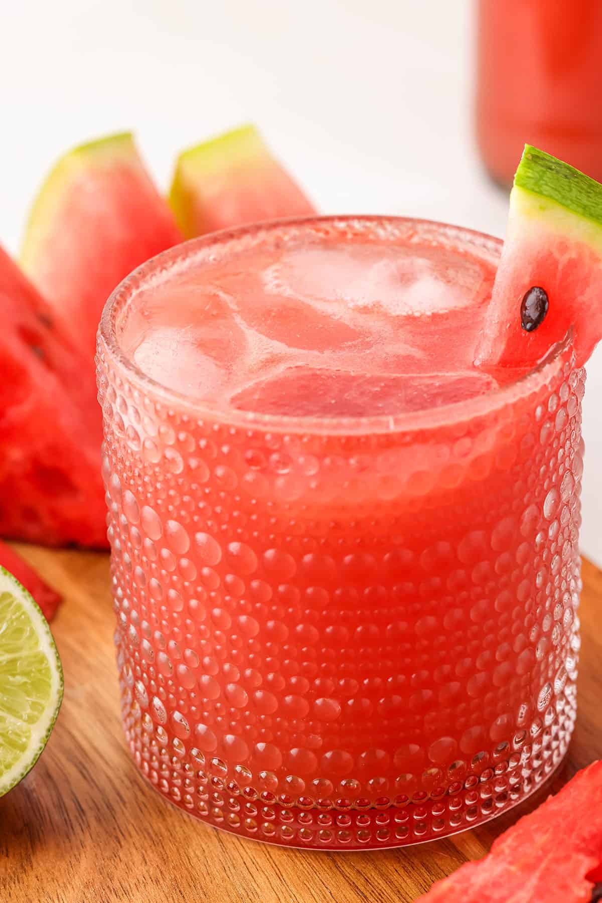 A glass of watermelon kombucha with ice garnished with a fresh slice of watermelon on the side of the glass, with watermelon and lime slices in the background. 