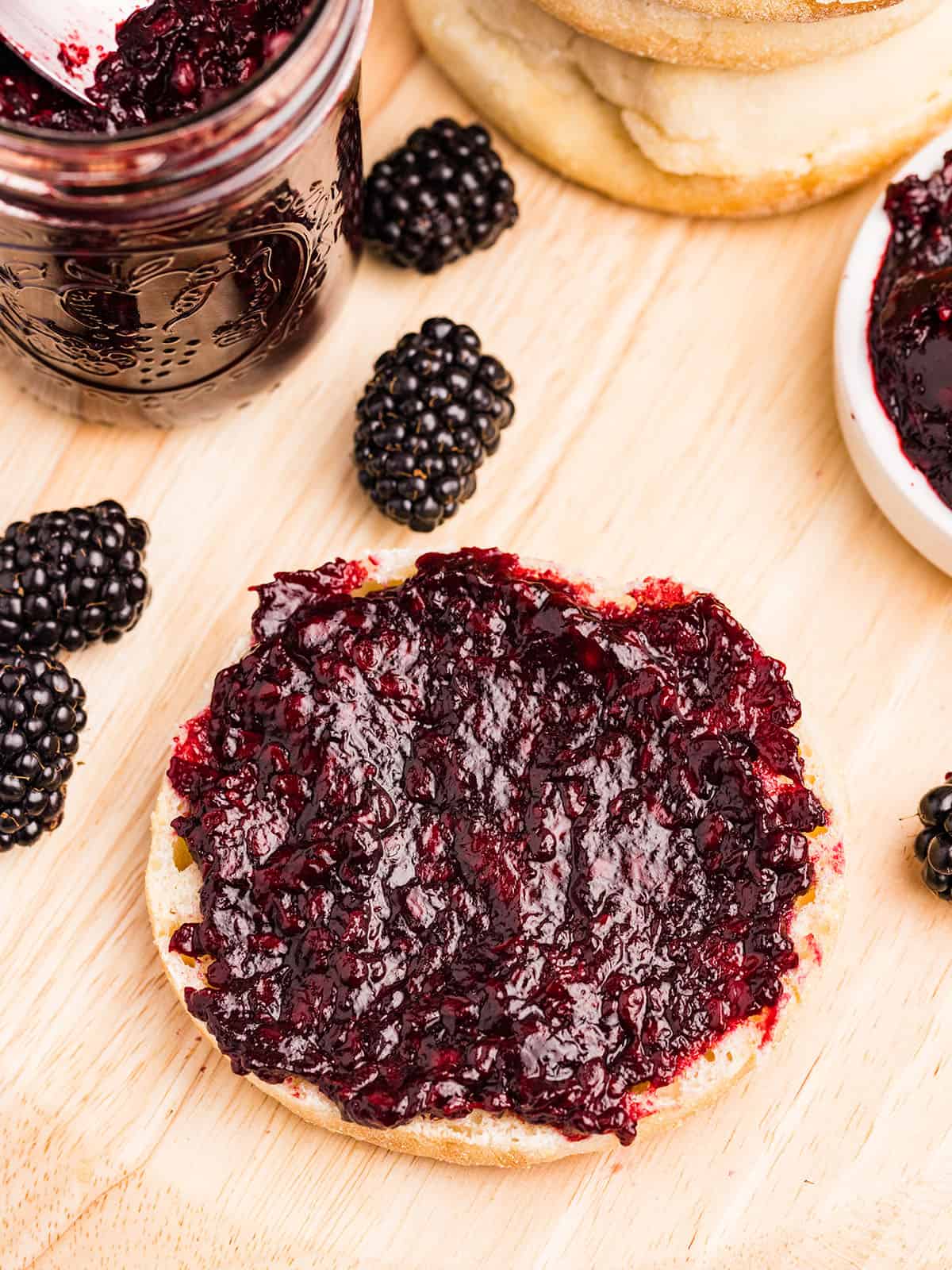 Blackberry jam spread on an English muffin, on a wood cutting board surrounded by fresh blackberries and a jar of blackberry jam. 