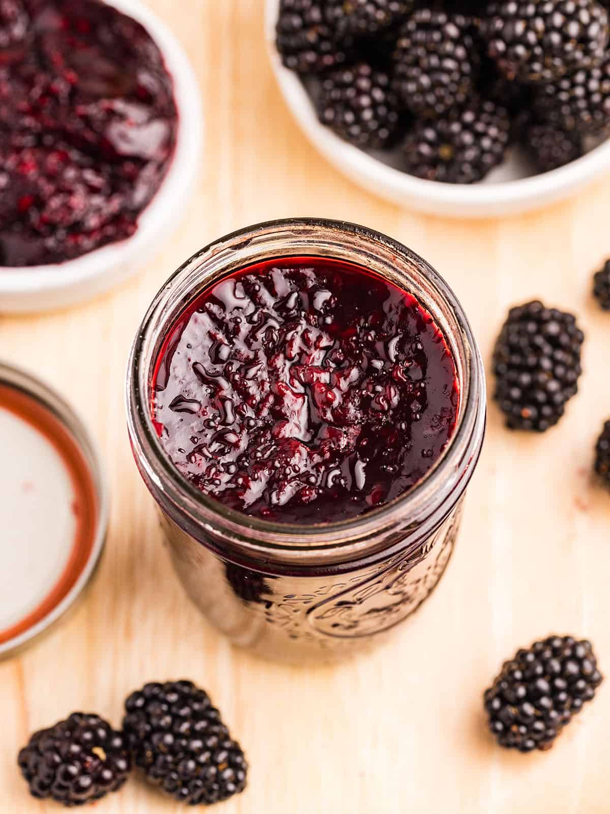 An open jar of blackberry jam on a wood cutting board surrounded by fresh blackberries. 