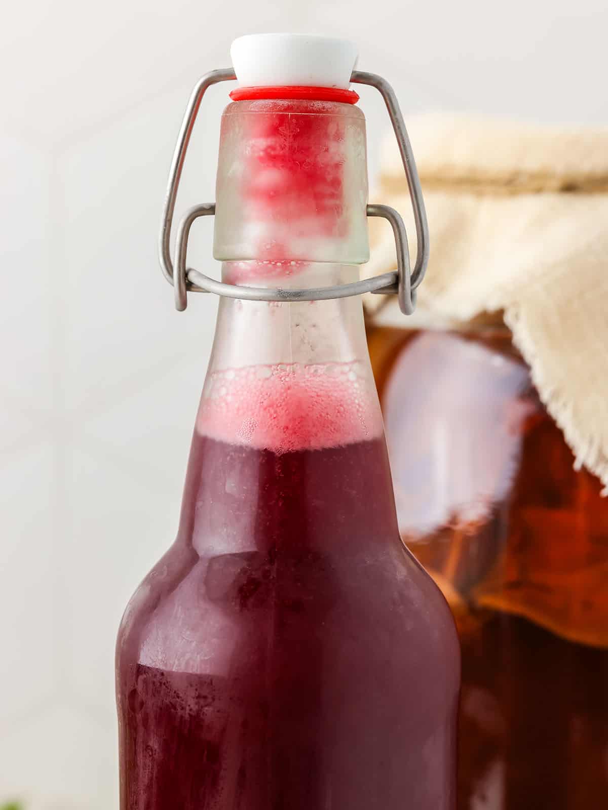 A close up of a closed flip top bottle of blackberry kombucha, with a large jar of first fermented kombucha in the background. 