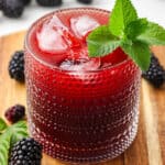 A collins glass of blackberry kombucha with ice, and a mint sprig garnishing. On a wood surface with fresh blackberries and mint surrounding.