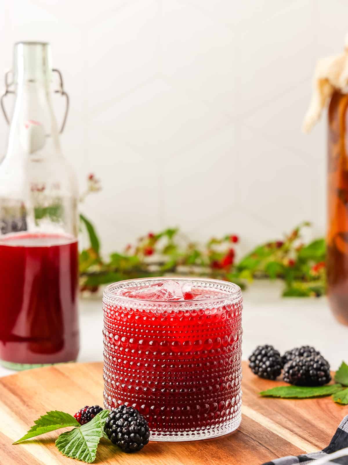 A glass of blackberry kombucha, on a wood surface surrounded by a half filled bottle, fresh mint and blackberries. 