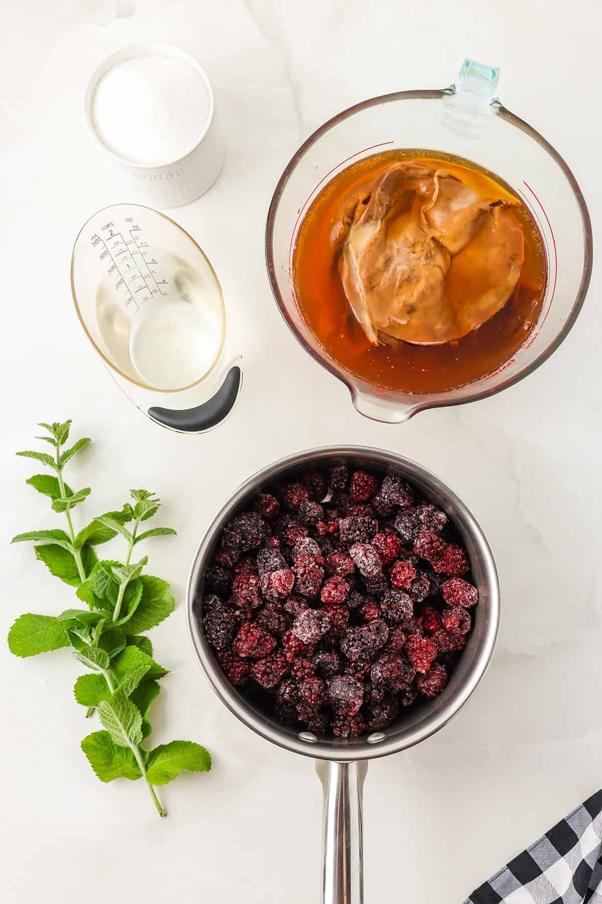 A white countertop with a pot of blackberries, a sprig of mint, water, and a bowl of kombucha, and a bowl of sugar. Top view. 