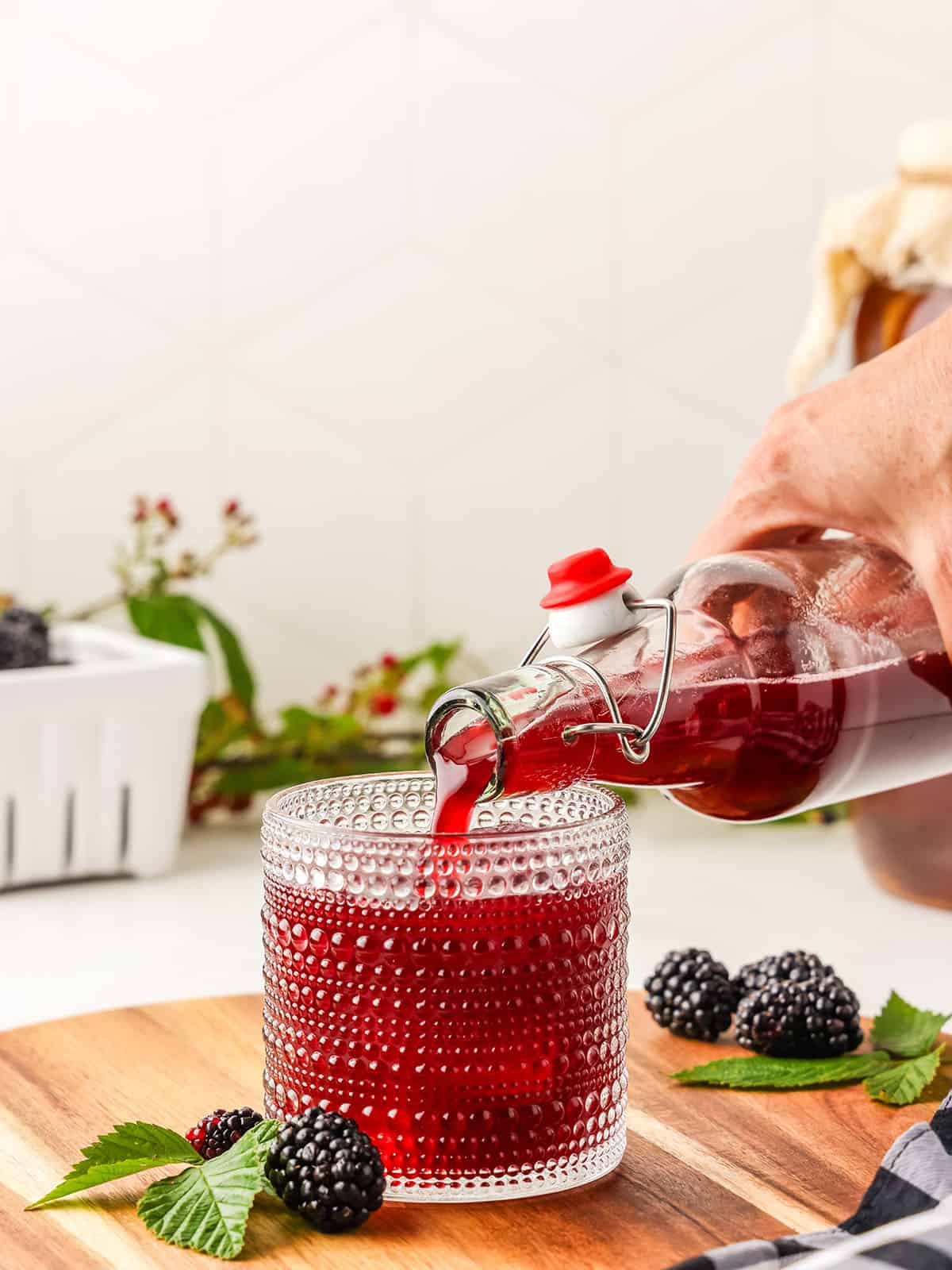 A bottle of blackberry kombucha pouring into a glass with ice, on a wood surface with a white background. Surrounded by fresh mint and blackberries. 