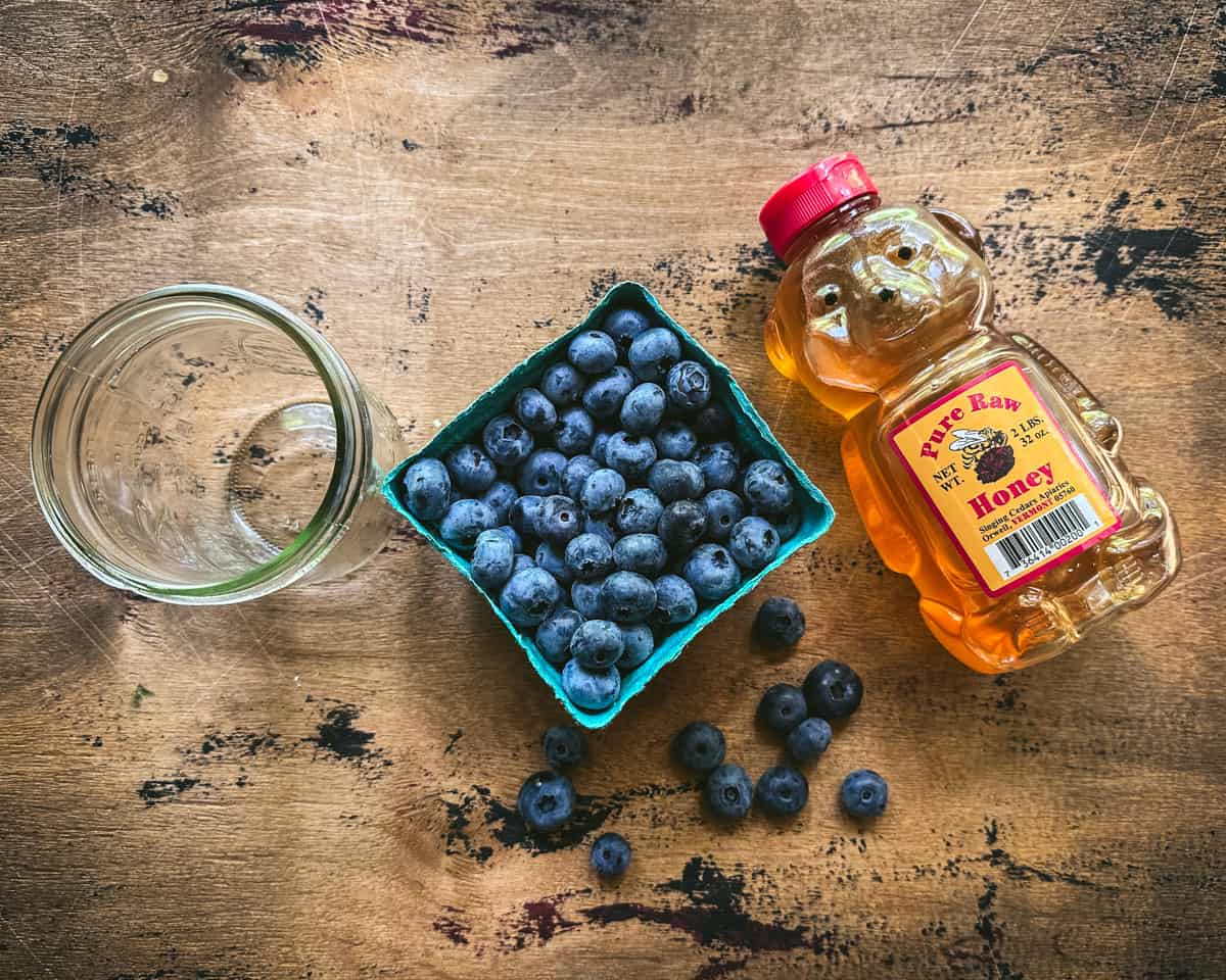 A honey bear, a pint of blueberries, and a jar on a wood surface, top view. 