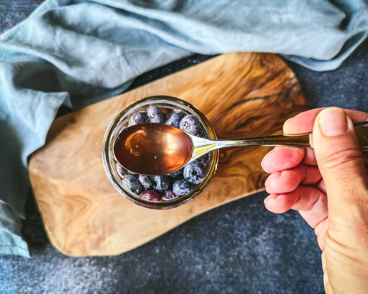 A jar of fermented honey blueberries with a spoon lifting up some honey, on a wood surface, top view. 