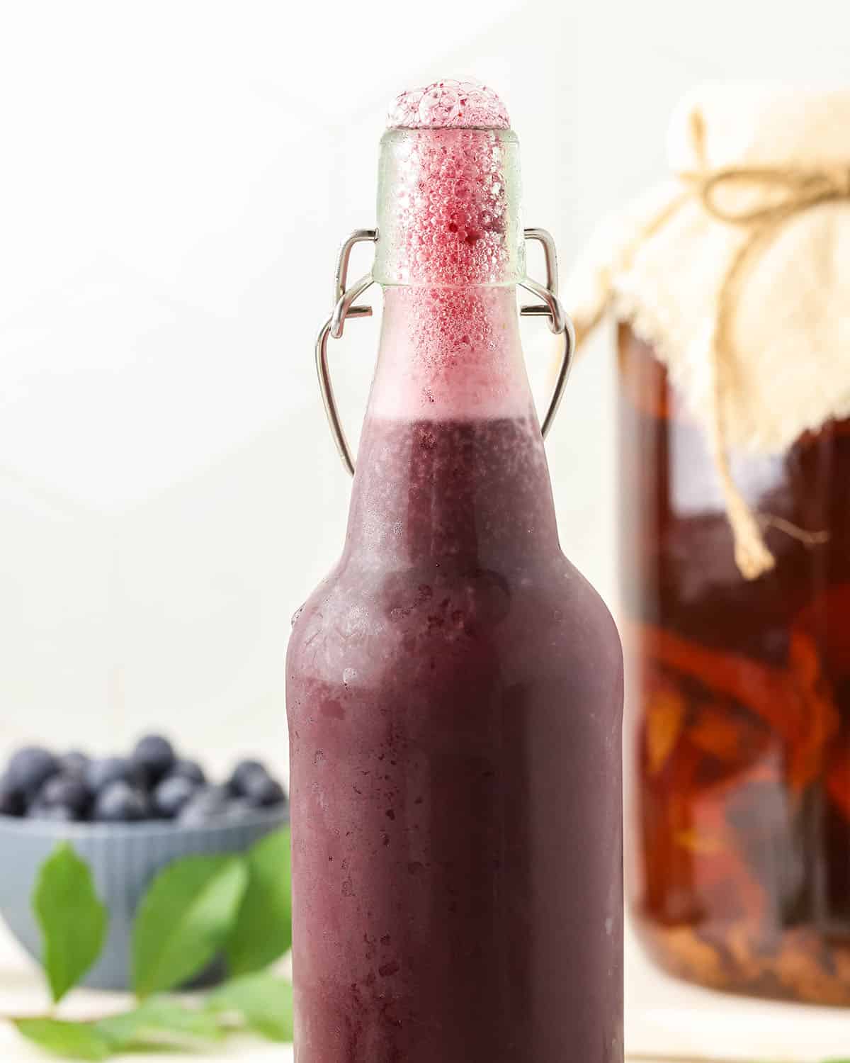 A bottle of fermented blueberry kombucha opened with bubbles fizzing to the top, with blueberries and kombucha in the background. 